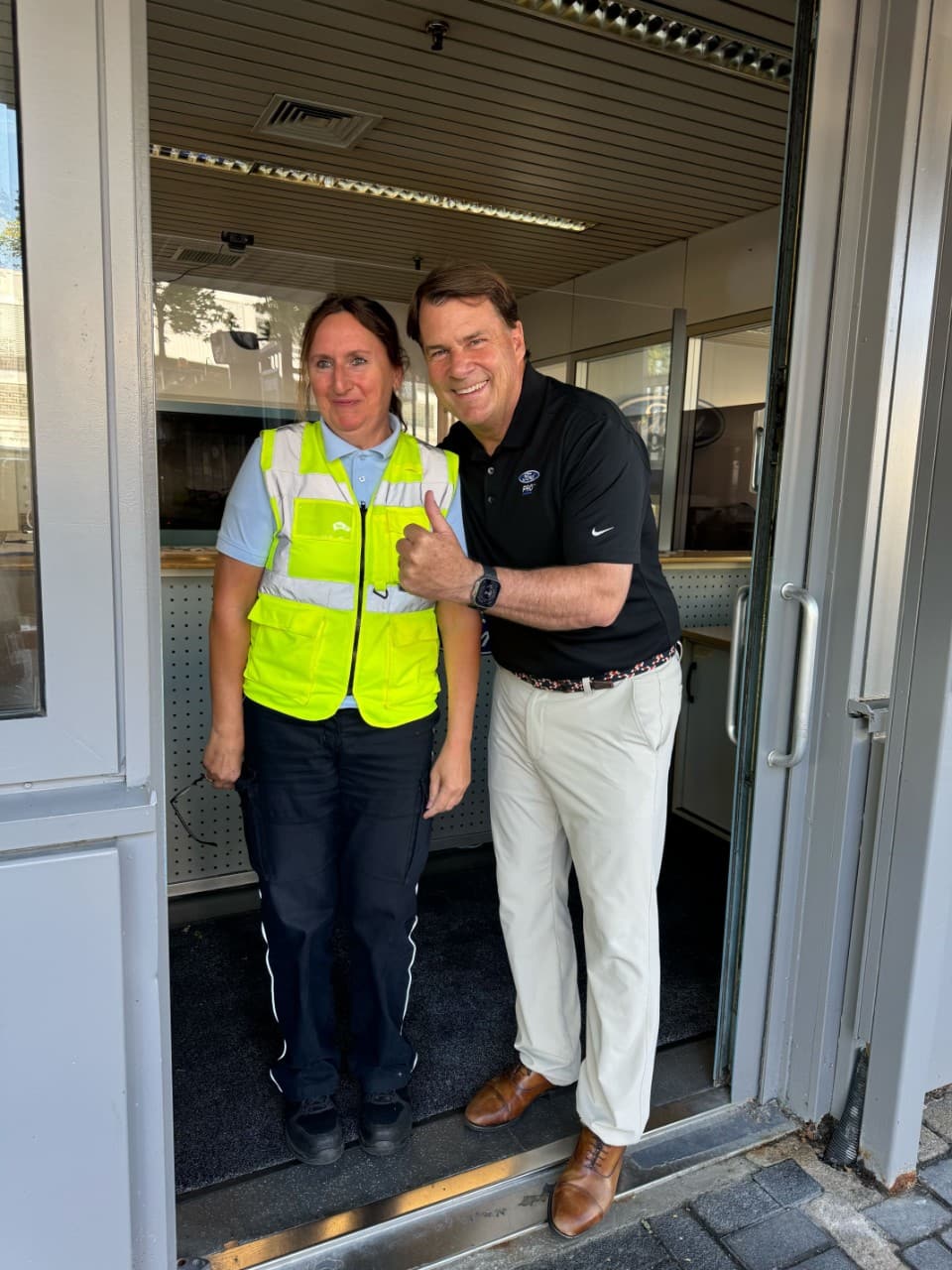 Jim Farley, dressed in khakis ad a dark blue polo, poses with a woman named Gabi Biermann, who wears a neon yellow vest.